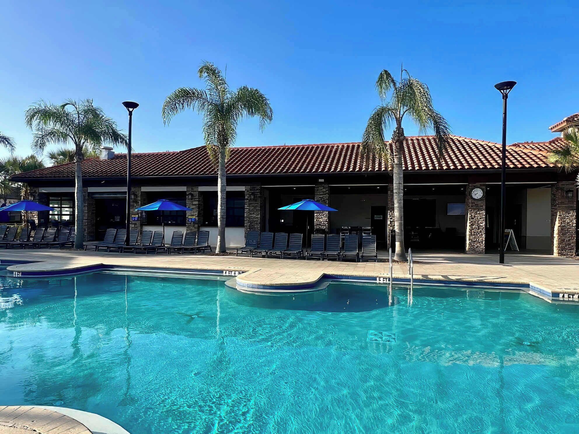 A pool area with lounge chairs and umbrellas, a building with a tiled roof, and palm trees.