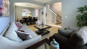 A modern living room with a sofa, armchair, and coffee table. There is a dining area with chairs and a table in the background. - Conservation View | Pool, Spa & Waterslides 4396 (Davenport)