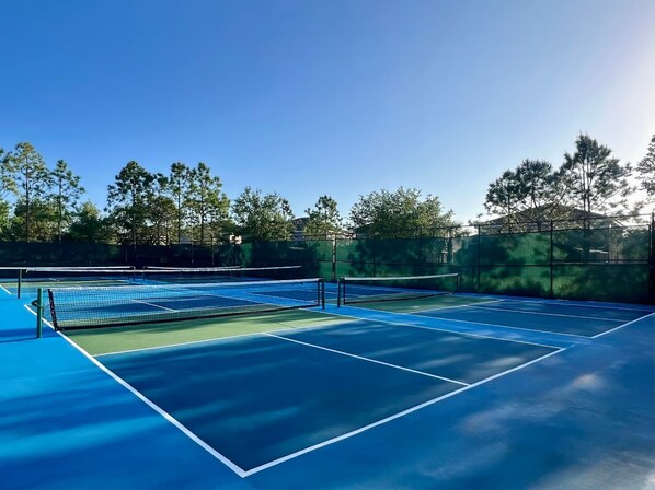 An outdoor tennis court with blue flooring and a net, surrounded by trees and a fence.