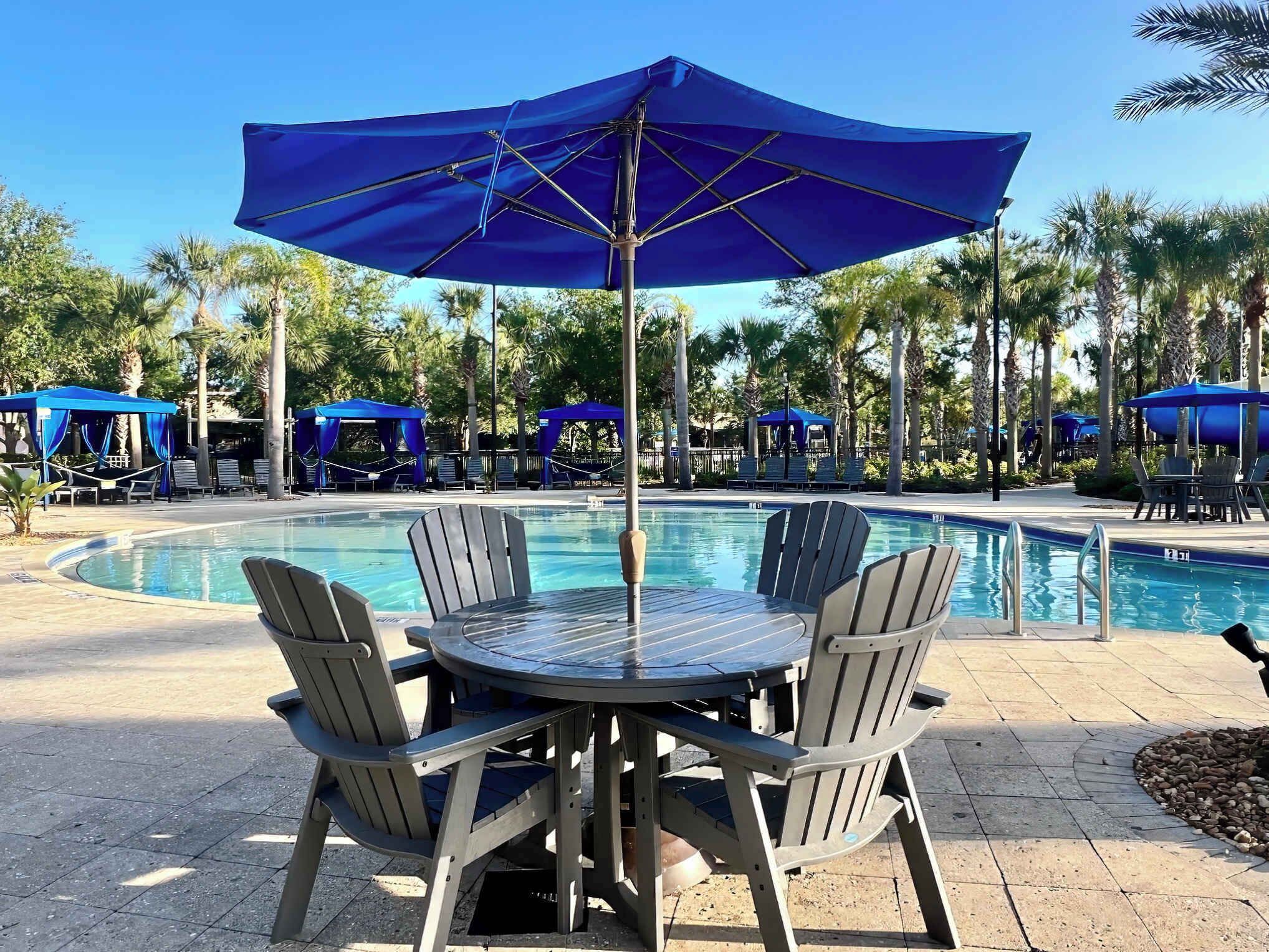Un espace aménagé au bord de la piscine, avec une table ronde et quatre chaises, entouré de parasols bleus et de chaises longues.