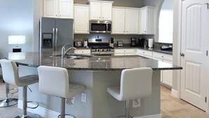 Una cocina moderna con gabinetes blancos, encimera de granito, electrodomésticos de acero inoxidable y una zona de bar con taburetes.