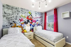 Un dormitorio con dos camas, un mural de Mickey Mouse, una ventana con cortinas rojas y una mesita de noche blanca.