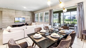 A modern living room with a sofa, dining table set for six, and a large window with curtains.