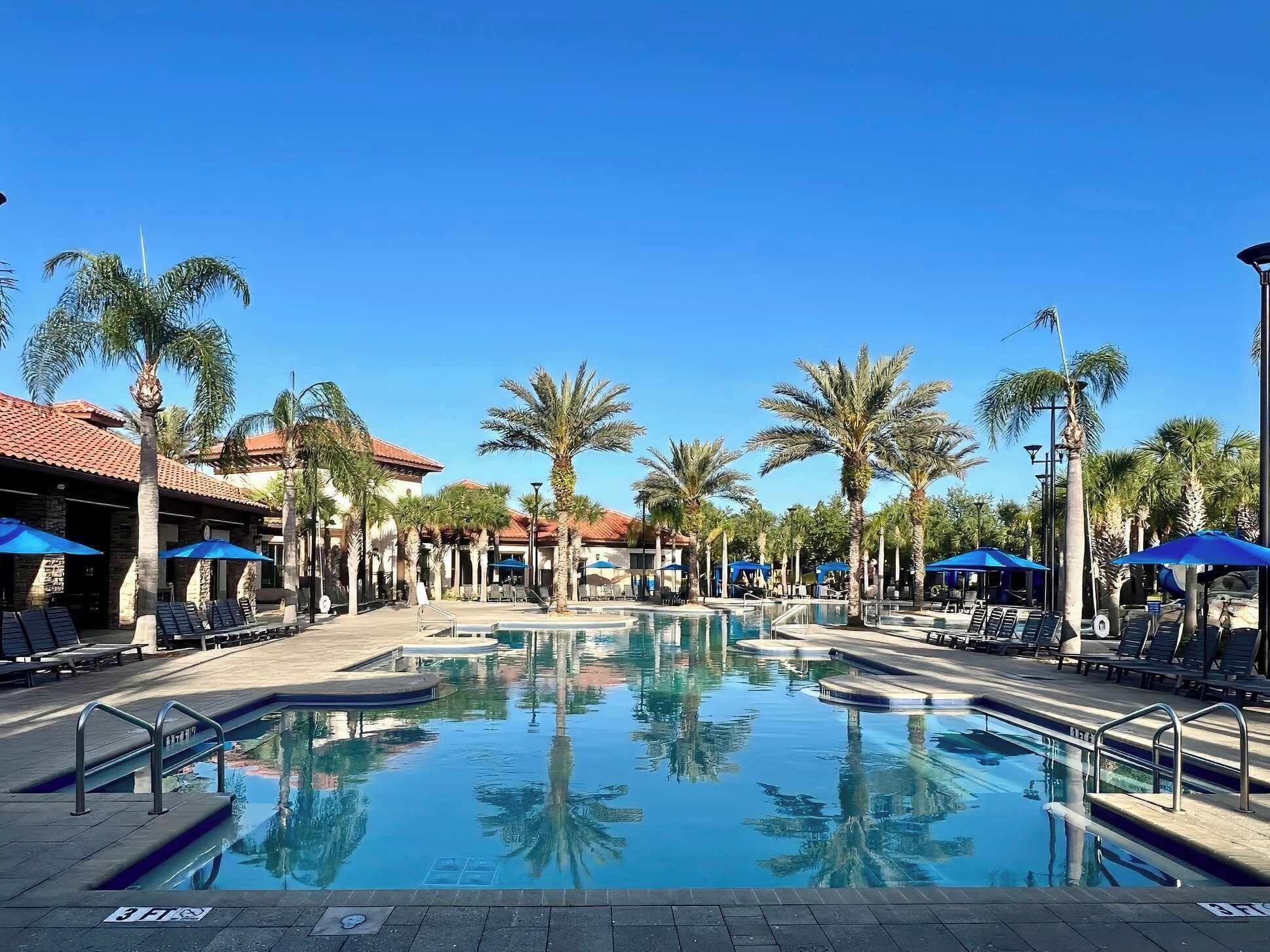 A pool area with lounge chairs and umbrellas, surrounded by palm trees and buildings with red-tiled roofs.