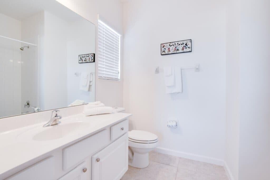 Une salle de bain moderne avec un lavabo blanc, un miroir et des toilettes. Des serviettes sont suspendues sur un porte-serviettes et un petit panneau décoratif est accroché au mur.