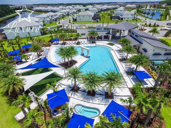 Una gran piscina rodeada de palmeras y toldos azules, con vistas a edificios residenciales al fondo.