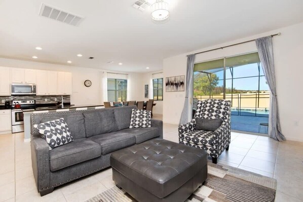 A modern living room with a grey sofa, a patterned armchair, and a leather ottoman. There is a kitchen area with white cabinets and a microwave. The room has large windows with curtains and a view of the outdoors.