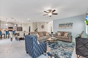 A modern living room with a sofa, coffee table, and patterned ottoman. There is a dining area with chairs and a table, and a kitchen in the background.