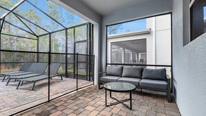 Une terrasse ensoleillée avec une véranda en verre, un canapé gris et une table ronde.