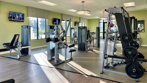 Ein Fitnessstudio mit verschiedenen Trainingsgeräten, einem Fernseher an der Wand und großen Fenstern für natürliches Licht.