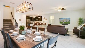 A modern dining area with a wooden table set for six, a kitchen in the background, and a living room with a sofa and chairs. - Game Room | No Rear Neighbors | 4 Miles Disney 2709 (Kissimmee)