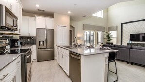 A modern kitchen with stainless steel appliances, a central island, and a grey sofa in the living area. - Splash Pool | Air Hockey | Lazy River | Disney 2755 (Kissimmee)