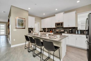 A modern kitchen with white cabinets, a central island with bar stools, a stainless steel refrigerator, and a microwave.