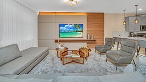 Una sala de estar moderna con un televisor de pantalla plana grande, sofás grises, una mesa de centro y una cocina con armarios grises y luces colgantes.