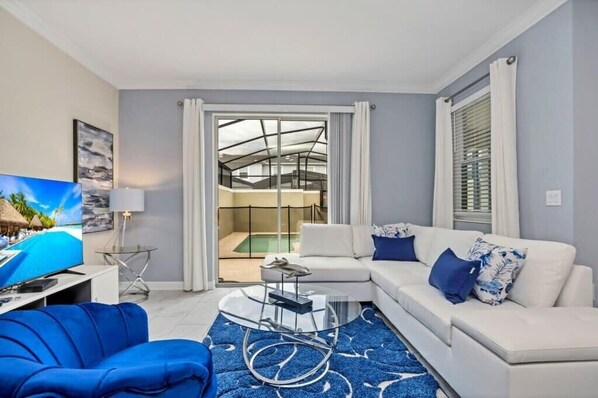 Un salon moderne avec un canapé blanc, un fauteuil bleu et une table basse en verre. Un téléviseur à écran plat est posé sur un meuble, et un balcon avec une porte vitrée donne accès à un espace extérieur.