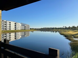 Un bâtiment moderne à étages multiples et doté de balcons, une étendue d’eau calme qui reflète la structure, et un ciel dégagé.