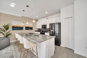 Una cocina moderna con armarios blancos, electrodomésticos de acero inoxidable, una isla central con taburetes de bar e iluminación colgante.
