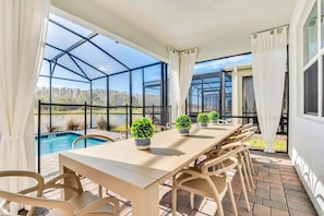 A covered patio with a table and chairs, overlooking a swimming pool and a forested area.