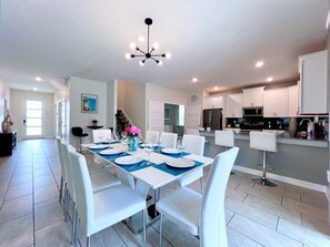 Une cuisine moderne avec des meubles blancs, un îlot central et un coin repas avec des chaises blanches et une nappe bleue.
