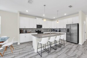 Cozinha moderna com armários brancos, geladeira de aço inoxidável, ilha de granito com banquetas e iluminação pendente.