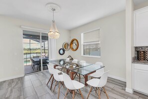 A dining area with a glass table, white chairs, a chandelier, and a view through glass doors.