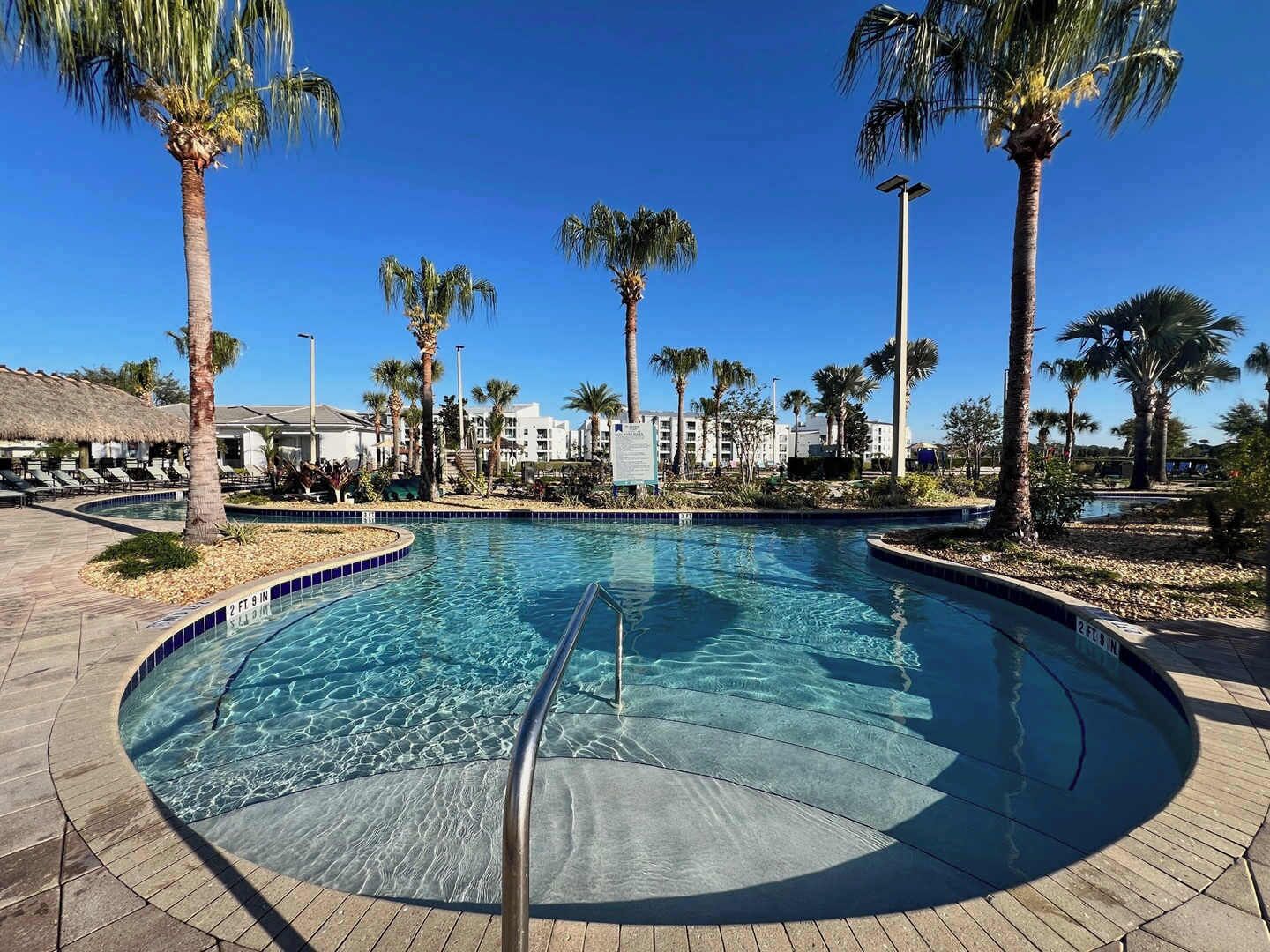 A clear swimming pool with a palm tree-lined deck, a thatched roof structure, and a poolside lounge area.