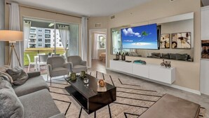 A modern living room with a large TV, comfortable seating, a white entertainment unit, and a view of the outdoors. - Great Value | Lazy River | 4 Miles to Disney 3180 (Kissimmee)