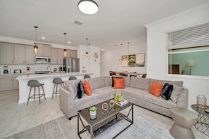A modern living room with a grey sofa, coffee table, and bar stools. There is a kitchen area with a microwave and refrigerator. - Splash Pool | Lazy River | Waterslides | Disney 4228 (Kissimmee)