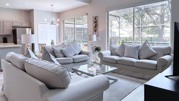 A modern living room with two sofas, a coffee table, and a TV. There is a kitchen in the background with cabinets and a refrigerator.