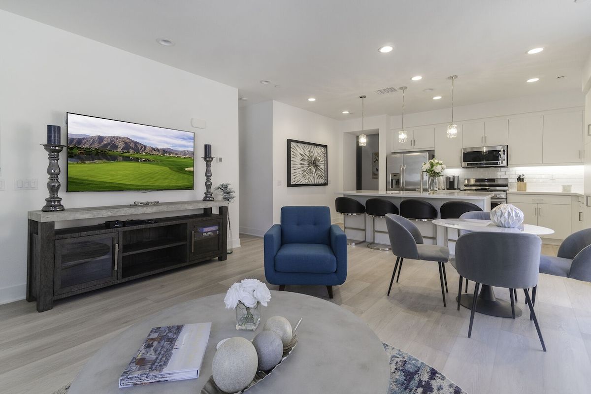 A modern living room with a flat-screen TV, a dining area with a round table, and a kitchen with white cabinets and stainless steel appliances.