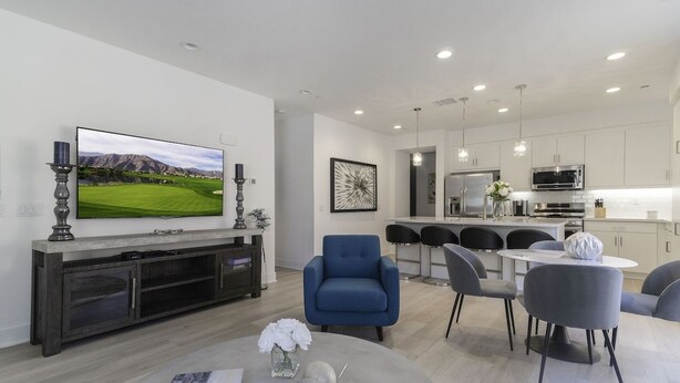 A modern living room with a flat-screen TV, a dining area with a round table, and a kitchen with white cabinets and stainless steel appliances. - Desert Serenity | Pool, Spa, BBQ, Near Courses (La Quinta)