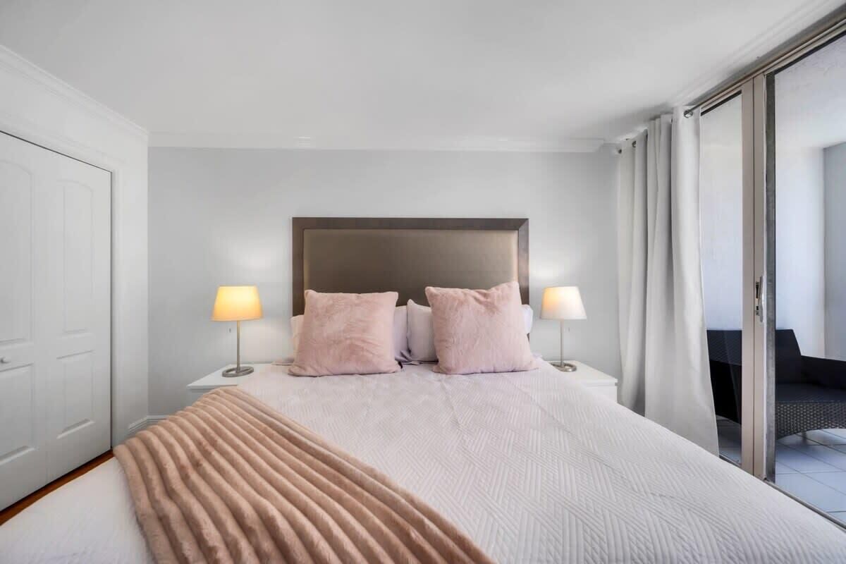 A modern hotel room with a large bed, two bedside lamps, a headboard, and a view of the outdoor area.