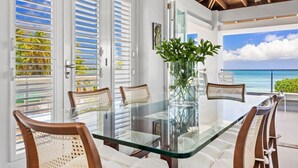 A glass-top dining table with wicker chairs, a vase with green plants, and a view of the ocean through a door with white shutters. - Casa Salinas | Spacious Waterfront Home with Pool (Cabo Rojo)