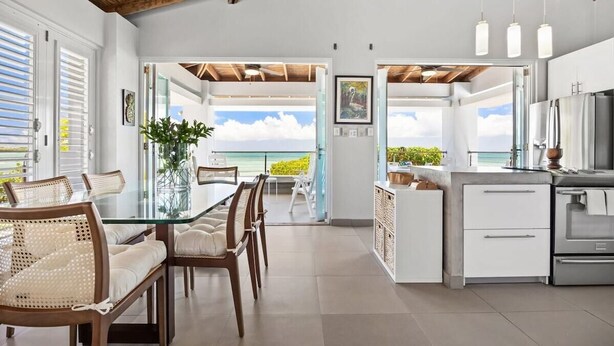 A modern kitchen with a view of the ocean, a dining area with a glass table, and a ceiling with exposed beams. - Casa Salinas | Spacious Waterfront Home with Pool (Cabo Rojo)