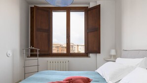 A bedroom with a bed, a radiator, a window with wooden shutters, and a view of buildings outside. - Siena: appartamento panoramico con terrazza, wi-fi e aria condizionata (Siena)