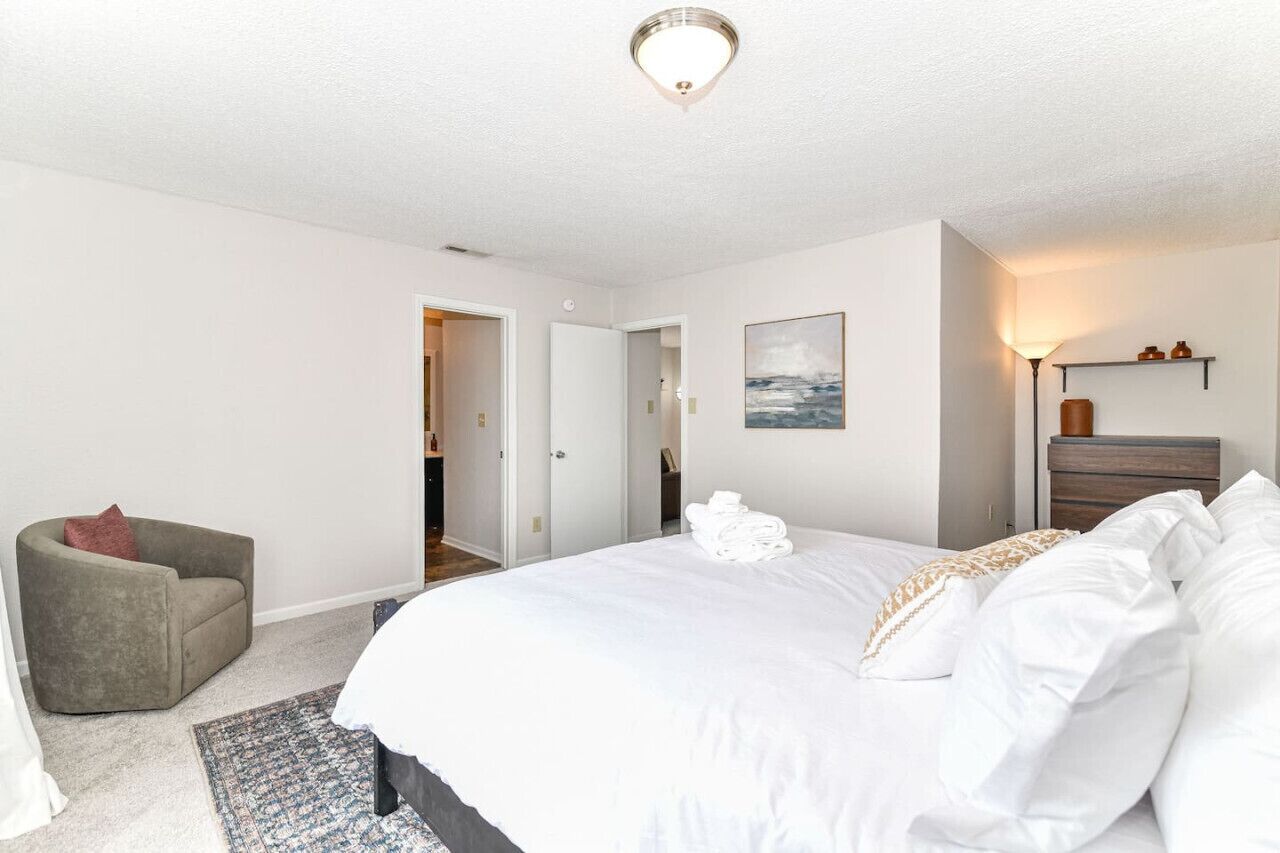 Ein Schlafzimmer mit einem Bett, einem Sessel, einem Teppich, einem Gemälde und einem Nachttisch mit Lampe.