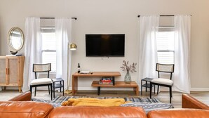 Un salon comprenant un téléviseur, deux fauteuils, une petite table avec des livres, un vase avec des fleurs et un canapé.
