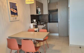 A compact kitchen with modern appliances, a dining area with a wooden table and orange chairs, and a wall-mounted artwork depicting a surfer. - Les Thermes 402 - Appt avec piscine commune (Concarneau)