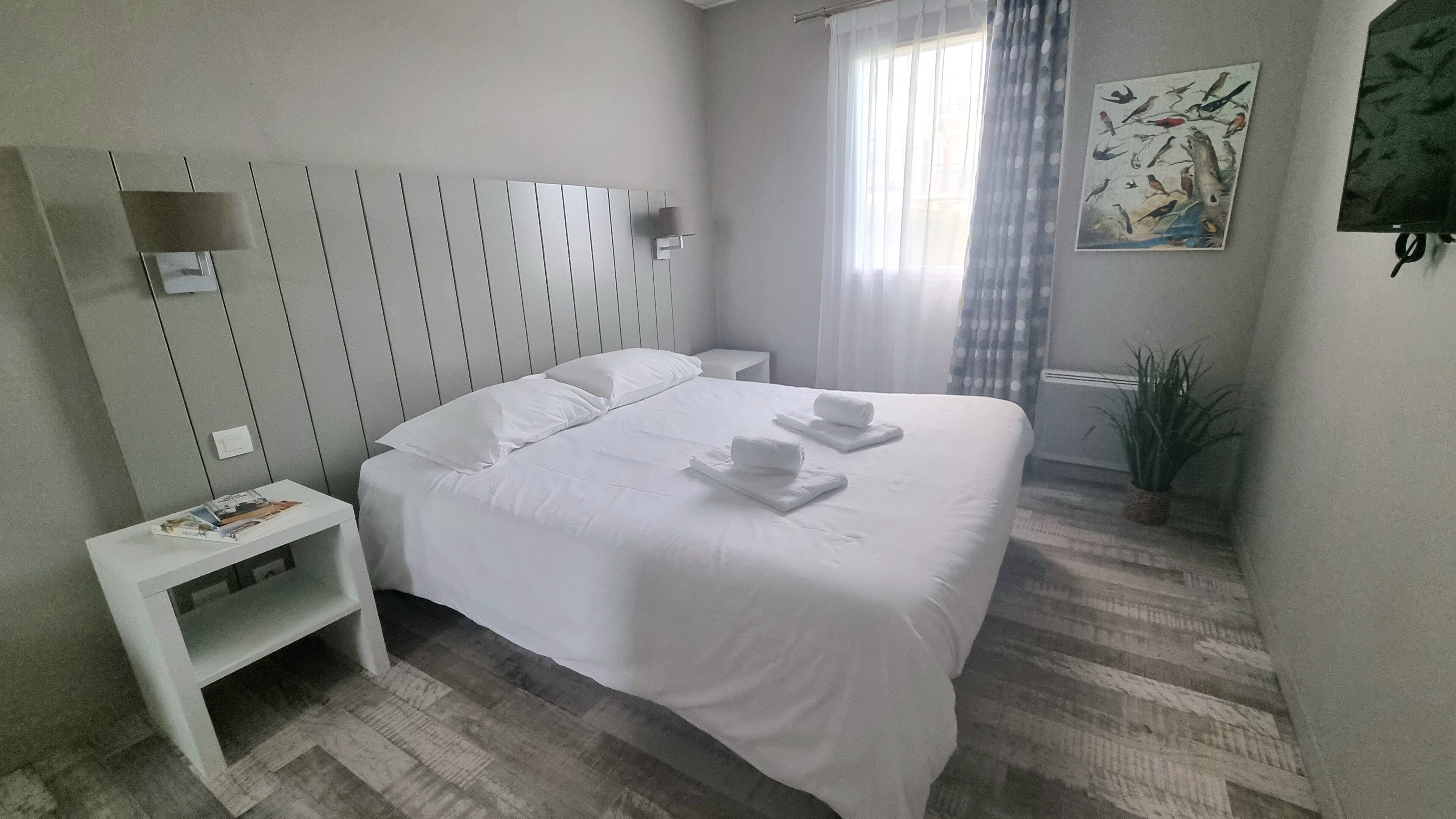 Ett hotellrum med en säng, ett nattduksbord, en liten hylla, ett fönster med gardiner och ett konstverk som hänger på väggen.