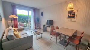 Ein modernes Wohnzimmer mit einer Couch, einem Couchtisch, einem Esstisch, Stühlen, einem Fernseher und einem großen Fenster mit Vorhängen.