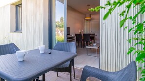 Un'area pranzo all'aperto con tavolo e sedie, una porta scorrevole in vetro che dà su una cucina e vista sul verde circostante.