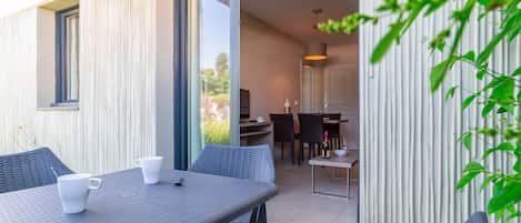 An outdoor dining area with a table and chairs, a sliding glass door leading to a kitchen, and a view of the surrounding greenery.