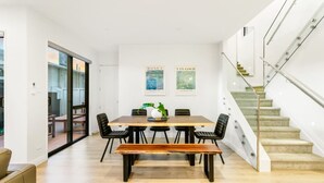 Ein moderner Essbereich mit einem Holztisch, schwarzen Stühlen und einer Bank. Zwei gerahmte Kunstwerke befinden sich an der Wand, und eine Treppe führt nach rechts.