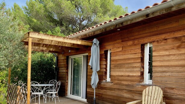 A wooden cabin with a covered patio, a table and chairs, and a single window. - Gite 4 Personnes Terrasse Jardin Piscine (aspiran)