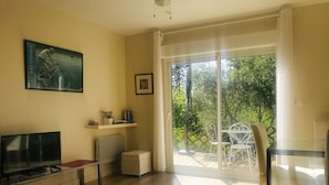 A living room with a flat-screen TV, a small table, a chair, a sofa, a potted plant, and a sliding glass door leading to an outdoor patio with a white metal table and chairs. - Gite 4 Personnes Terrasse Jardin Piscine (aspiran)