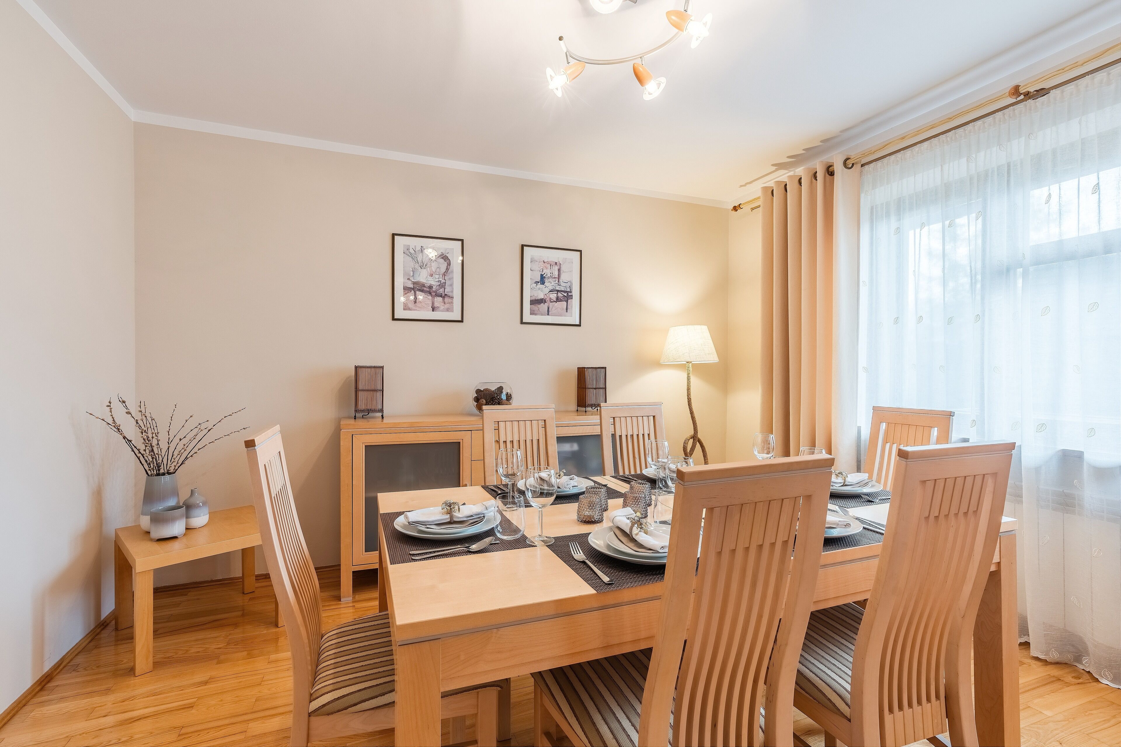Un coin repas avec une table en bois dressée pour quatre personnes, entourée de chaises, une petite table d’appoint avec un vase, et des œuvres d’art encadrées accrochées au mur.