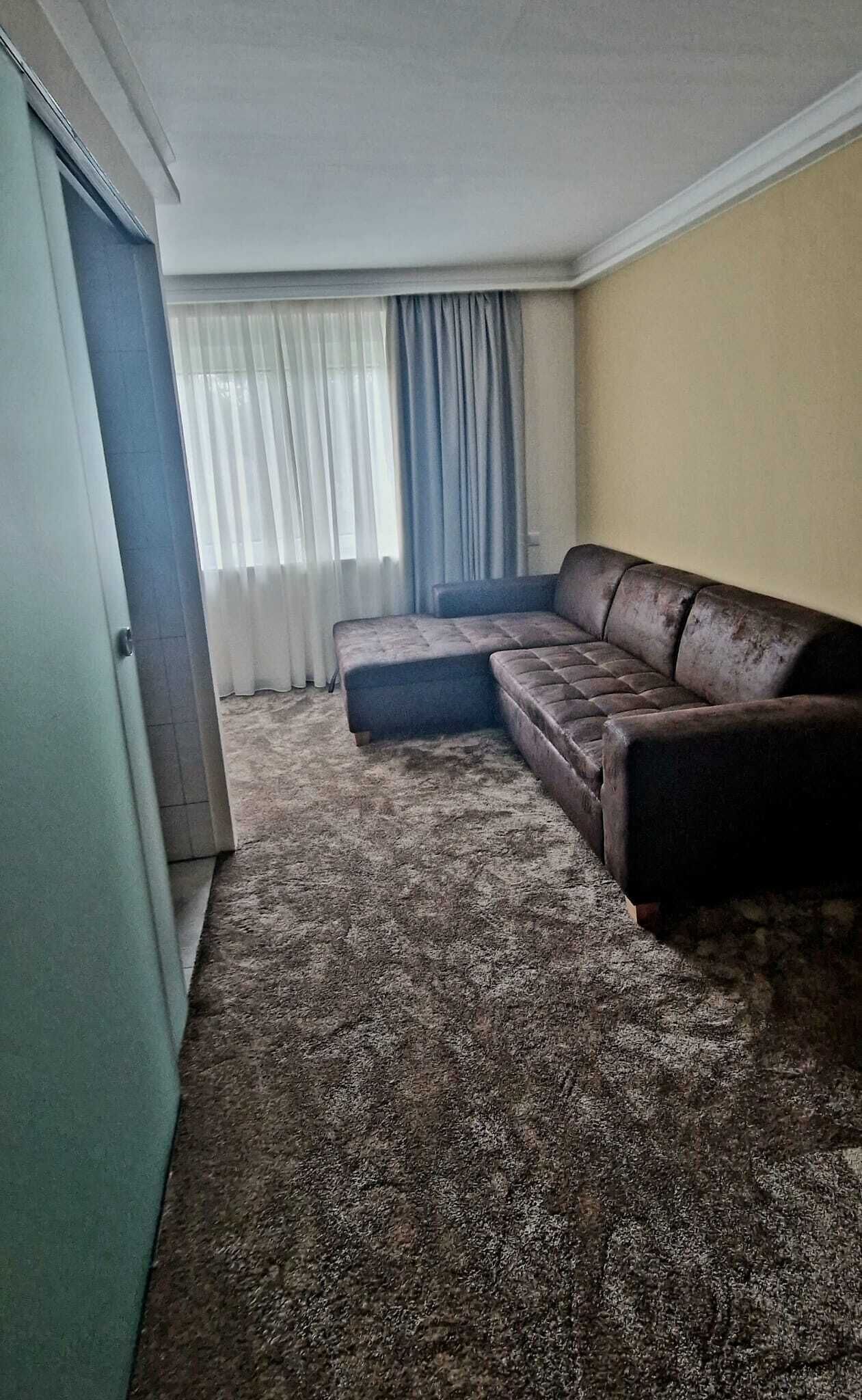 Ein Wohnzimmer mit braunem Ledersofa, einem gemusterten Teppich, einer Tür und einem Fenster mit Vorhängen.