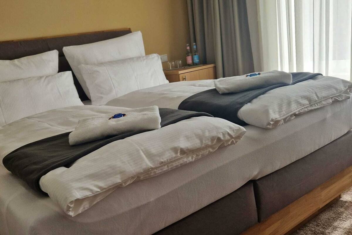 Un lit bien fait, avec une literie blanche et grise, une tête de lit en bois et une table de chevet sur laquelle se trouvent une bouteille et des verres.
