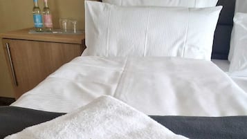Una cama bien hecha con sábanas blancas, un cabecero gris y una mesita de noche con dos botellas y dos vasos.