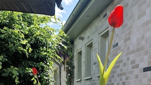 Un patio al aire libre con mesas y sillas de madera, dos jarrones blancos con flores rojas y un edificio de ladrillo al fondo.